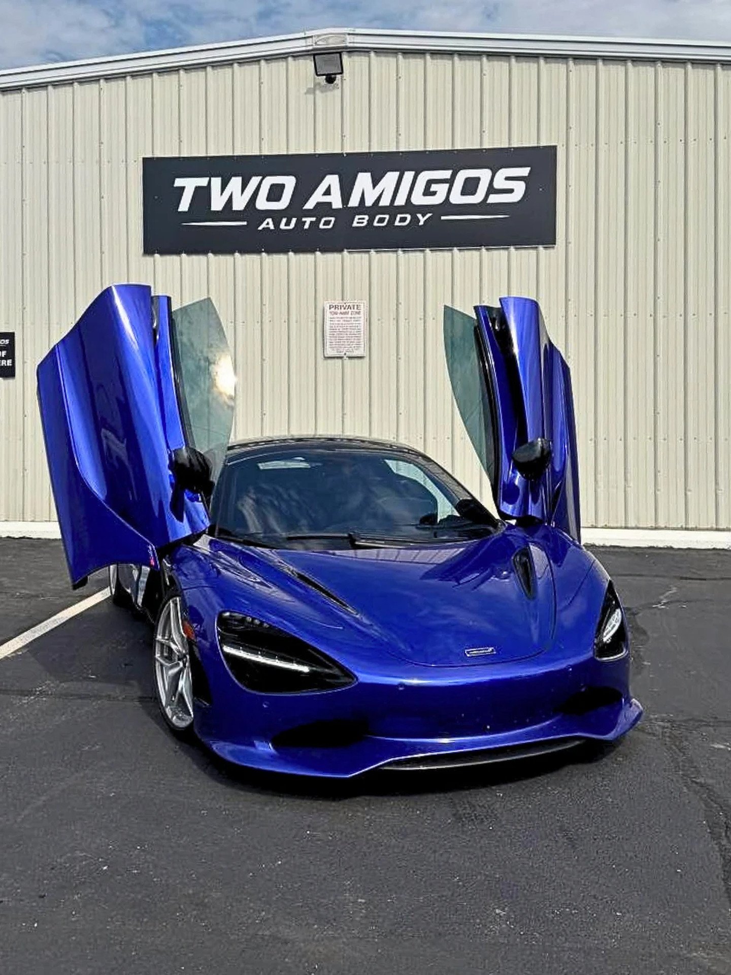 Blue McLaren 750S with both dihedral doors raised, parked in front of the Two Amigos Auto Body sign